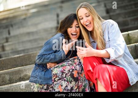 Deux jeunes femmes à la recherche à un drôle de chose sur leur téléphone intelligent à l'extérieur, assis sur les marches. Les filles d'amis à rire. Banque D'Images