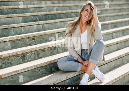 Belle jeune femme blonde souriante sur les marches urbaines. Belle jeune femme caucasienne souriant en milieu urbain. Fille blonde portant des vêtements décontractés dans la rue. Femme avec une veste élégante et un Jean bleu assis dans les escaliers. Banque D'Images