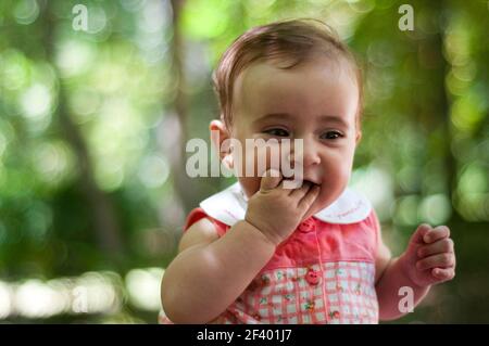 Bébé fille de six mois s'amusant dehors. Portrait en gros plan d'une petite fille de six mois avec sa main dans la bouche et son arrière-plan défoqué. Banque D'Images