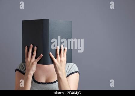 Jeune femme avec son visage caché derrière un livre noir qu'elle tient avec les deux mains, ouvert, la lisant. Portrait avec arrière-plan gris et espace de copie. Banque D'Images