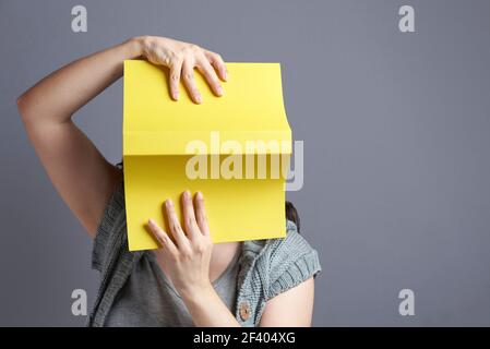 Jeune femme avec son visage caché derrière un livre jaune qu'elle tient ouvert, tourné, avec les deux mains. Portrait avec arrière-plan gris et espace de copie. Conc Banque D'Images