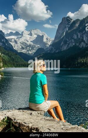 Fille blonde assise sur le rocher en profitant de la vue sur les montagnes majestueuses et l'eau turquoise cristal à Gosau See, Autriche, glacier de Dachstein en arrière-plan. Banque D'Images