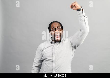 Un gars afro-américain enthousiaste portant une chemise élégante et décontractée célébrant la victoire, levant le poing serré, hurlant oui heureux, un gars noir hurlant ecstatic, concept de triomphe, studio tourné isolé sur gris Banque D'Images