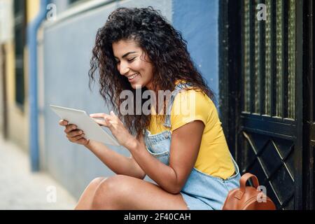 Jeune femme arabe en utilisant sa tablette numérique à l'extérieur. L'Afrique du Nord en souriant les tenues avec black curly hairstyle. Banque D'Images