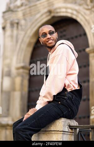 Jeune homme noir portant des vêtements et des lunettes de soleil, assis sur l'arrière-plan. Jeune africain guy avec les salopettes en plein air Banque D'Images