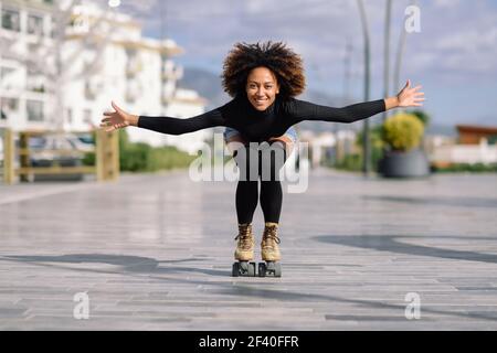 Jeune femme noire monter à cheval sur patins à l'extérieur sur la rue urbain à bras ouverts. Smiling girl avec coiffure afro à roues alignées sur un jour ensoleillé