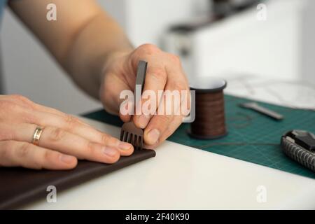 Hommes main tenant un marteau à sculpter en cuir et un poinçon de trou de burin en acier et fait un portefeuille en cuir dans son atelier. Processus de travail avec un marron Banque D'Images