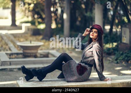 Jeune belle femme portant un manteau d'hiver et une casquette assis sur un banc dans un parc urbain. Style de vie et mode. Jeune belle fille portant un manteau d'hiver et une casquette assis sur un banc dans un parc urbain. Banque D'Images