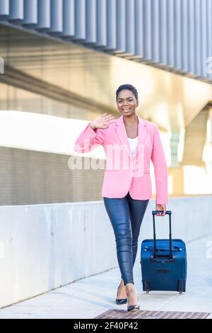 Une femme d'affaires afro-américaine se trouve dans une valise à roulettes près du bâtiment des bureaux d'affaires. Femme noire marchant avec un sac de voyage portant une veste rose. Femme noire marchant avec un sac de voyage portant une veste rose. Banque D'Images