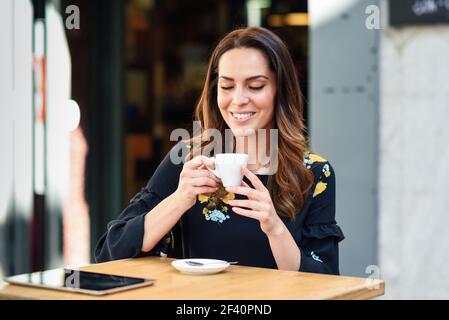 Belle femme buvant un café dans un café-bar urbain. Jeune femme assise à table sur une terrasse extérieure. Fille avec la californie met en vedette la coiffure.. Femme d'âge moyen qui boit du café dans un café-bar urbain. Banque D'Images