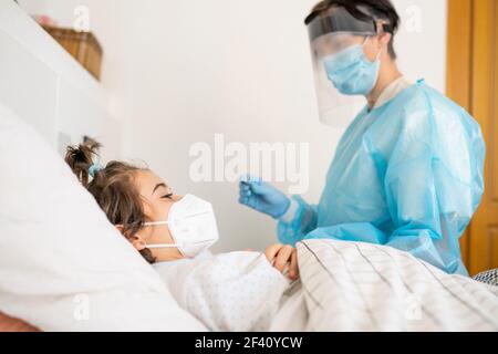 Médecin protégé par un équipement de protection personnelle examinant une petite fille avec un stéthoscope à la maison. Médecin examinant une petite fille avec un stéthoscope à la maison. Banque D'Images