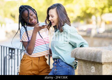 Deux amies en plein air enregistrent une note vocale avec un smartphone. Amis multiethniques... Deux amies qui s'amusent ensemble dans la rue. Amis multiethniques. Banque D'Images