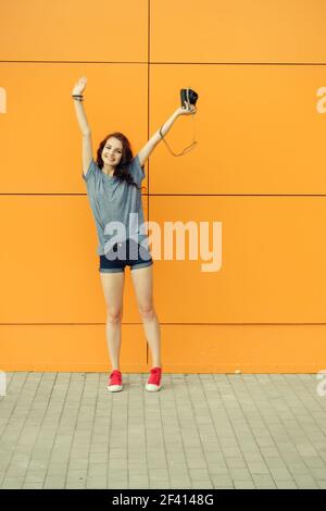 Hipster fille avec appareil photo vintage dans une main lever ses mains devant le mur orange, espace de copie, image de tons vintage. Hipster fille avec photo vintage dans une main lever ses mains devant le mur orange, espace de copie, image tonifiée Banque D'Images