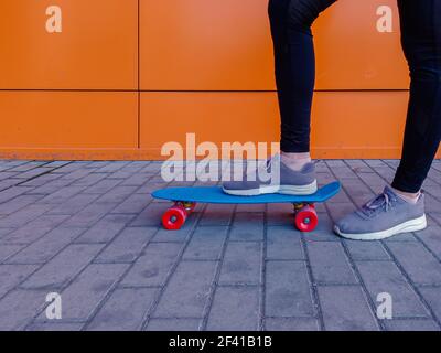 Photo d'une jeune fille qui a mis son pied dans des baskets sur un skateboard contre un mur orange vif. Photo d'une fille qui a mis son pied dans des baskets sur un skateboard. Banque D'Images