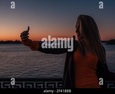 Jolie jeune femme en tricot tenant le chandail prend selfie sur la rue de la ville la nuit devant le coucher du soleil de la rivière. Une jolie tenue femelle prend le selfie dans la rue de la ville la nuit, devant le coucher du soleil sur la rivière Banque D'Images