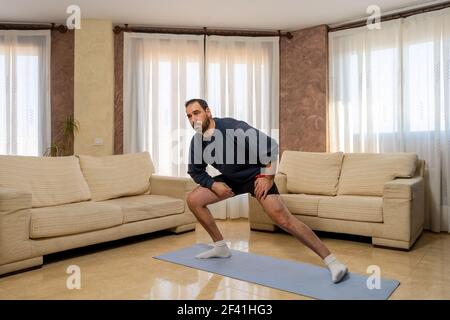 Placez l'homme barbu dans des vêtements de sport sombres en faisant des étirements ravisseurs dans le salon à la maison. Concept d'exercice Banque D'Images
