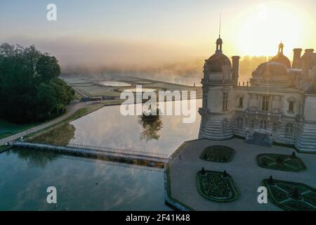 Vue du matin sur la surface de l'eau et les jardins du château dans la brume. Elève la vue contre le soleil levant, silhouette du château de Chantilly, Oise, France. Banque D'Images