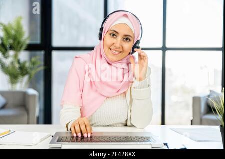 Femme arabe souriante dans le hijab, assistance à la clientèle au bureau, assis avec un casque, tapant sur le clavier en touchant l'écouteur avec la main, il n'y a pas de requête qu'elle ne peut pas résoudre, aimer le travail Banque D'Images