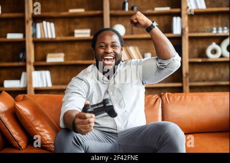 L'homme d'inspiration afro-américaine noire exprime la joie après avoir gagné le jeu, assis sur un grand canapé en cuir, jouant avec le joystick dans les jeux vidéo en ligne, s'amusant seul à la maison, en utilisant la console de jeux Banque D'Images