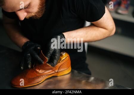 Vue rapprochée du cordonnier avec des gants noirs peignant les chaussures en cuir brun clair avec les doigts. Banque D'Images