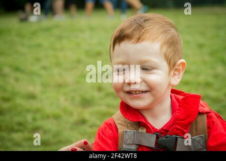 Portrait d'un joli bébé à tête rouge aux yeux bleus souriant portant une veste rouge dans un parc. Banque D'Images