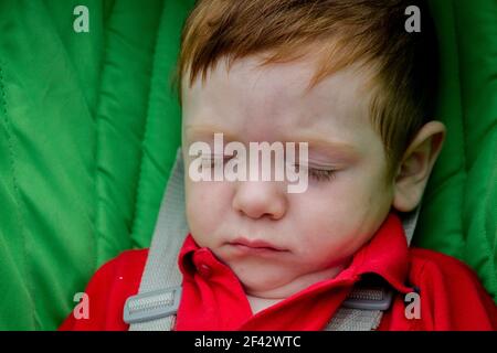 Portrait d'un adorable bébé à tête rouge portant une chemise rouge et dormant dans une poussette verte Banque D'Images