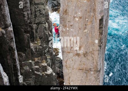 L'homme aventureux monte sur une colonne de roche exposée avec des falaises marines et l'océan en arrière-plan dans le Totem Pole, Cape Hauy, Tasman National Park, Tasmanie, Australie. Banque D'Images