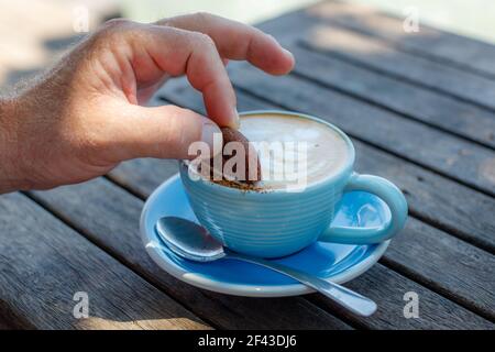 Un homme plongeant un cookie dans une tasse bleue de cappuccino sur une table en bois dans un café. Avec espace. Banque D'Images