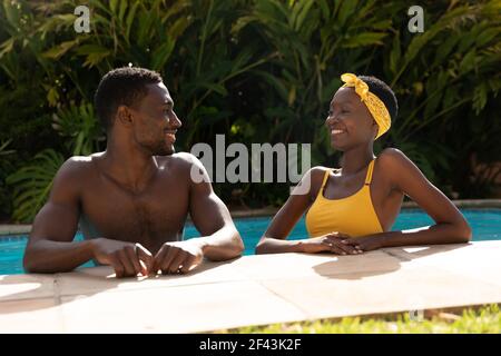 Couple afro-américain dans l'eau penchée au bord de la piscine par temps ensoleillé terrasse dans le jardin Banque D'Images
