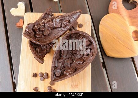 Gros plan de l'œuf de Pâques au chocolat amer avec des amandes croustillantes sur une planche de bois. Entouré de noix de Saint-Jacques au chocolat et de coeurs en bois. Banque D'Images