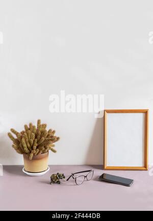 Sur une table contre un mur blanc se trouve un cadre photo en bois et une fleur de cactus en pot. Il y a des lunettes et un smartphone à côté. Vue de face, espace de copie Banque D'Images