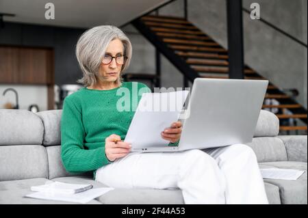Une femme d'affaires mûre et occupée à porter des lunettes et un pull vert assis sur le canapé avec un ordinateur portable sur ses genoux, lisant et vérifiant des documents, étudiant sous contrat ou travaillant à distance en ligne depuis la maison Banque D'Images