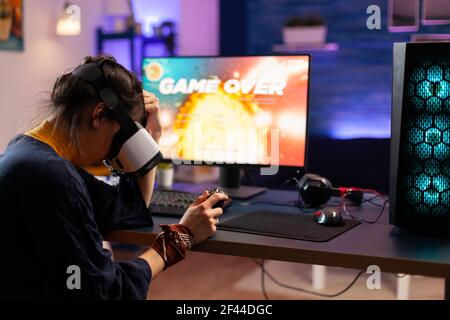 Joueur en ligne perdant la compétition de tir avec des écouteurs de réalité virtuelle. Joueur vaincu utilisant une manette professionnelle pour le championnat assis sur une chaise de jeu tard dans la nuit dans la salle de séjour Banque D'Images