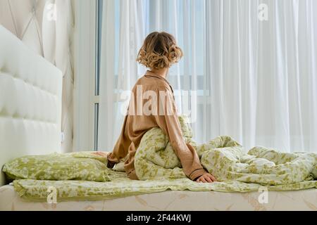 Vue arrière de la jeune femme qui s'étire les bras assis sur le lit dans la chambre à coucher donnant sur la fenêtre Banque D'Images