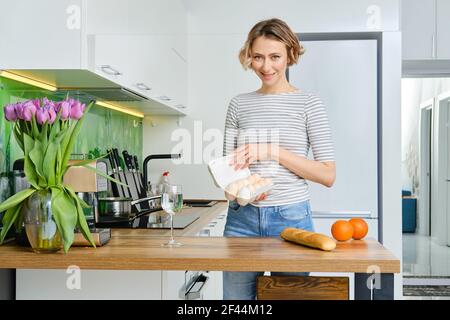 Une jeune femme positive prend des œufs hors de la boîte pour petit déjeuner Banque D'Images