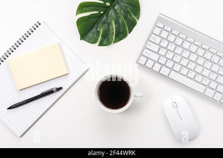 Espace de travail avec clavier d'ordinateur, fournitures de bureau, feuille verte et tasse à café sur fond blanc. Banque D'Images