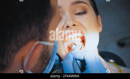 dentiste dans un masque médical utilisant un foret dentaire tout en traitant les dents de la femme, flou de premier plan Banque D'Images