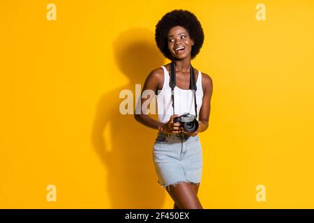 Portrait d'une jeune fille à cheveux ondulés plutôt gaie et mince tenant entre les mains digicam pour des souvenirs isolés sur fond jaune vif Banque D'Images