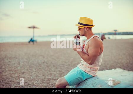 Jeune homme sportif élégant en vêtements brillants, chapeau de paille et lunettes de soleil, assis sur la mer, boire un cocktail rafraîchissant et apprécier ses vacances. Banque D'Images