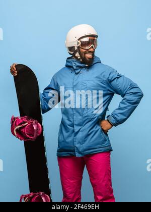 Snowboardeur mâle avec surf des neiges en costume de ski brillant isolé sur arrière-plan bleu Banque D'Images