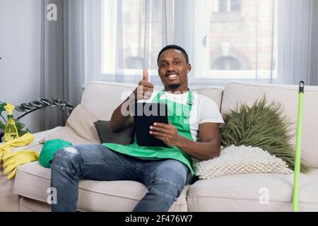 Concierge afro-américain souriant assis sur un canapé avec une tablette et montrant pouce vers le haut Banque D'Images