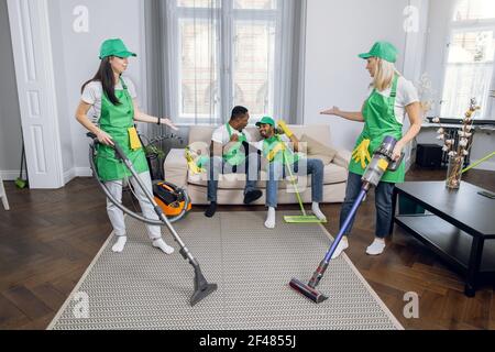 Deux femmes confuses dans le nettoyage uniforme appartement moderne tandis que deux hommes multiraciaux assis sur le canapé et jouant des jeux sur la tablette. Concept de personnes et de technologie. Banque D'Images