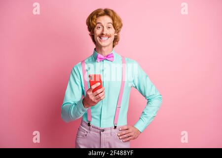 Photo de jeune funky positif funky drôle homme porte suspender noeud papillon avec smartphone isolé sur fond rose Banque D'Images