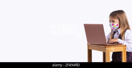 Bannière,format long.Une écolière de race blanche en uniforme et un masque médical s'assoit à une table avec un ordinateur portable.Le concept de formation pendant Covid 19 Banque D'Images