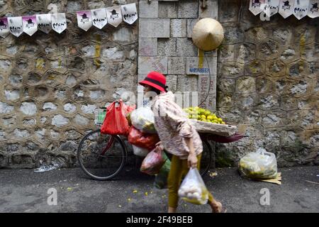 Hanoï, Vietnam - septembre 2015 : femme au chapeau rouge portant des sacs de shopping sur le marché de la rue. Vieux vélo avec des oranges derrière le mur Banque D'Images