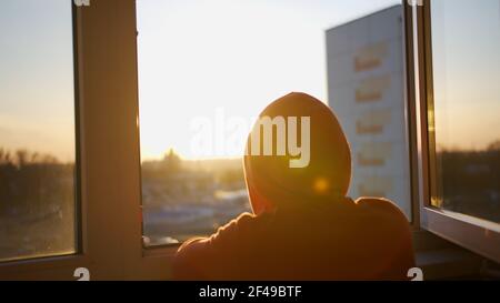 Un gars dans un sweat à capuche avec une capuche sur son la tête regarde le coucher du soleil en se tenant debout à l'air libre fenêtre Banque D'Images