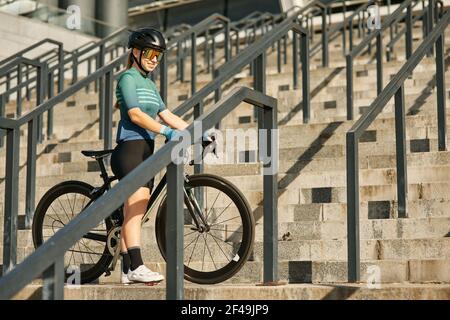 Jeune cycliste professionnelle gaie en vêtements de cyclisme et équipement de protection souriant, debout sur les marches avec son vélo pendant son entraînement à l'extérieur Banque D'Images
