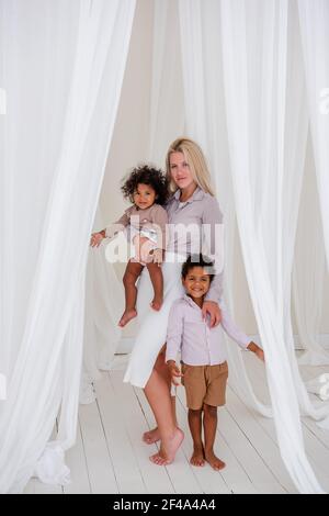 Une jeune mère blanche blonde détient des enfants afro-américains dans les armes. Femme embrasse, épouse fille et fils parmi les coupes blanches de tulles envolées. Papillon Banque D'Images