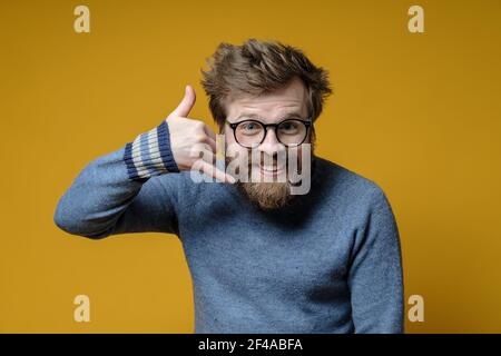 Drôle shaggy-cheveux, barbu homme portant des lunettes et un pull faire un geste de main m'appeler et regarde l'appareil photo. Arrière-plan jaune. Banque D'Images