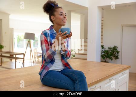 Femme de race mixte assise sur le plan de travail buvant un café et souriant dans une cuisine Banque D'Images